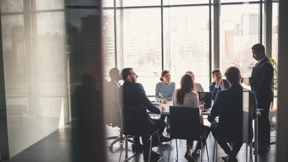 Seven people in business attire sit around a conference table in a modern office with large windows, engaged in a meeting with a Pittsburgh Bankruptcy Lawyer to discuss financial restructuring options.