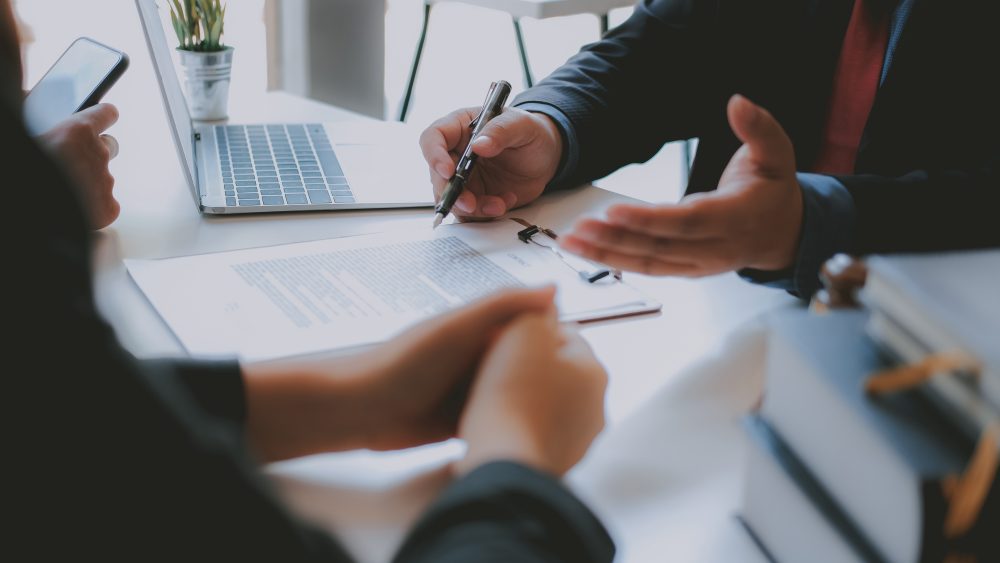 Two people sit at a table discussing a document with a clipboard, laptop, and stack of folders visible—perhaps consulting with a Pittsburgh Bankruptcy Lawyer as one gestures while the other holds a pen.