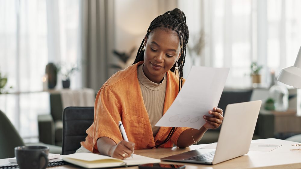 Woman sitting at a desk with a laptop, holding a paper and writing in a notebook in a modern, well-lit office—perfect for a busy Pittsburgh Bankruptcy Lawyer working on client cases.