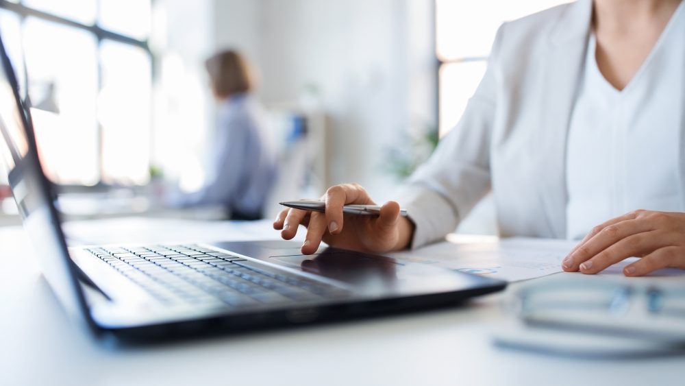 A person in business attire works at a desk with a laptop, holding a pen, with documents and glasses nearby; another person is blurred in the background.