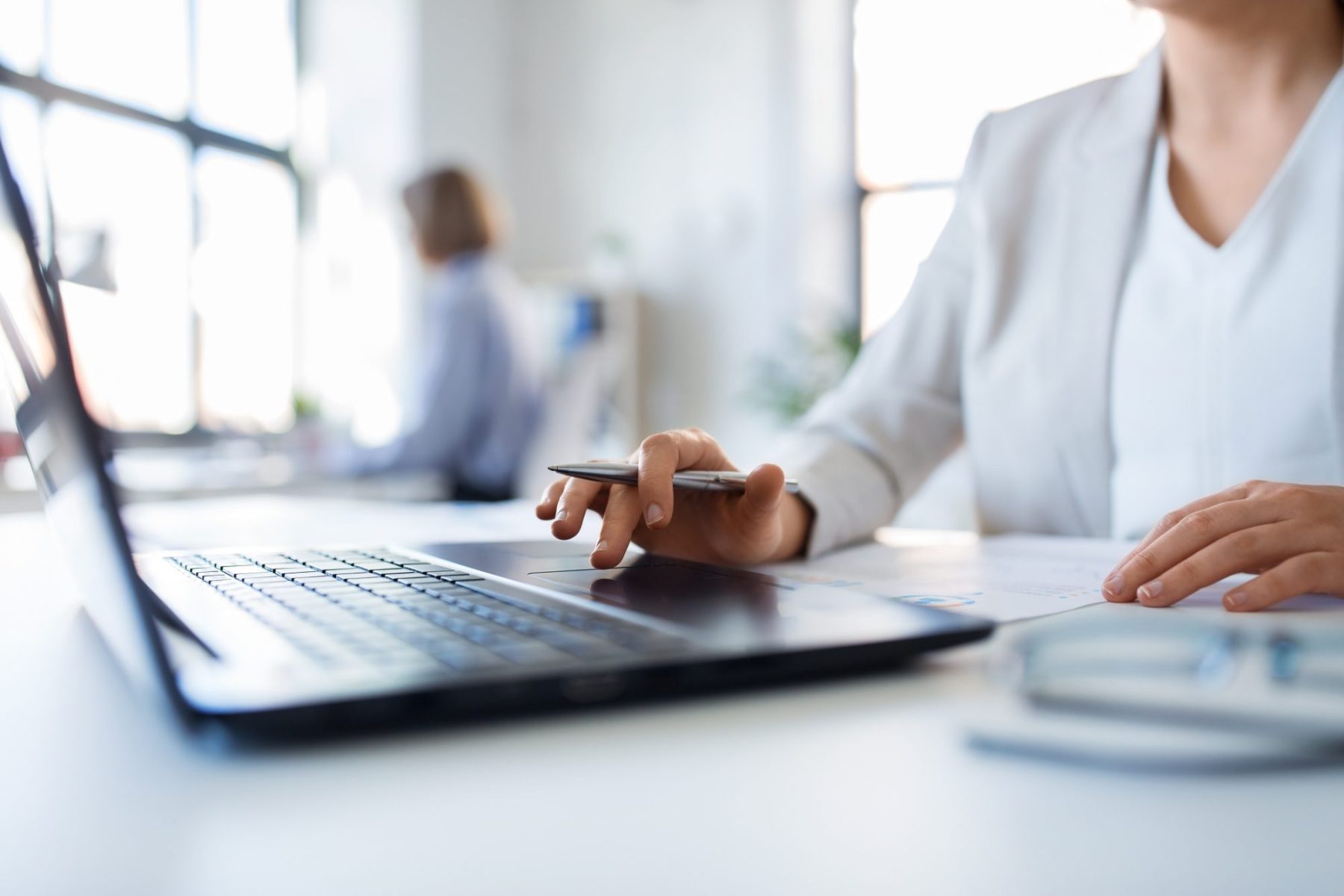 A person in business attire works at a desk with a laptop, holding a pen, with documents and glasses nearby; another person is blurred in the background.