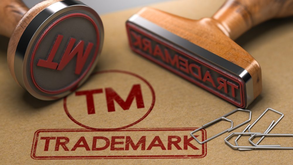 Two wooden rubber stamps—one upright, one tilted—plus three paperclips rest on a brown surface with a red "TM TRADEMARK" stamp, symbolizing protection from a Pittsburgh trademark law attorney.