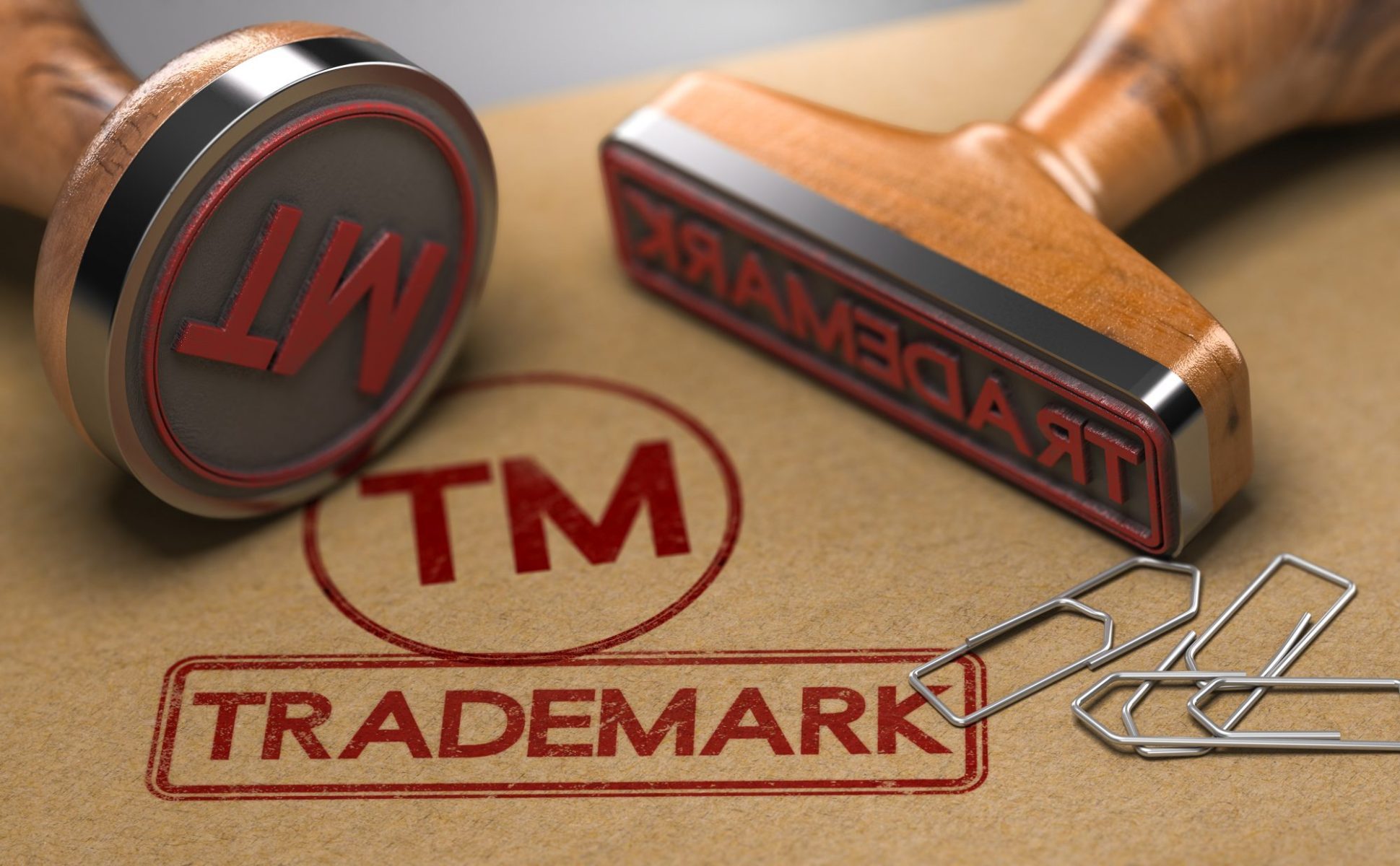 Two wooden rubber stamps—one upright, one tilted—plus three paperclips rest on a brown surface with a red "TM TRADEMARK" stamp, symbolizing protection from a Pittsburgh trademark law attorney.