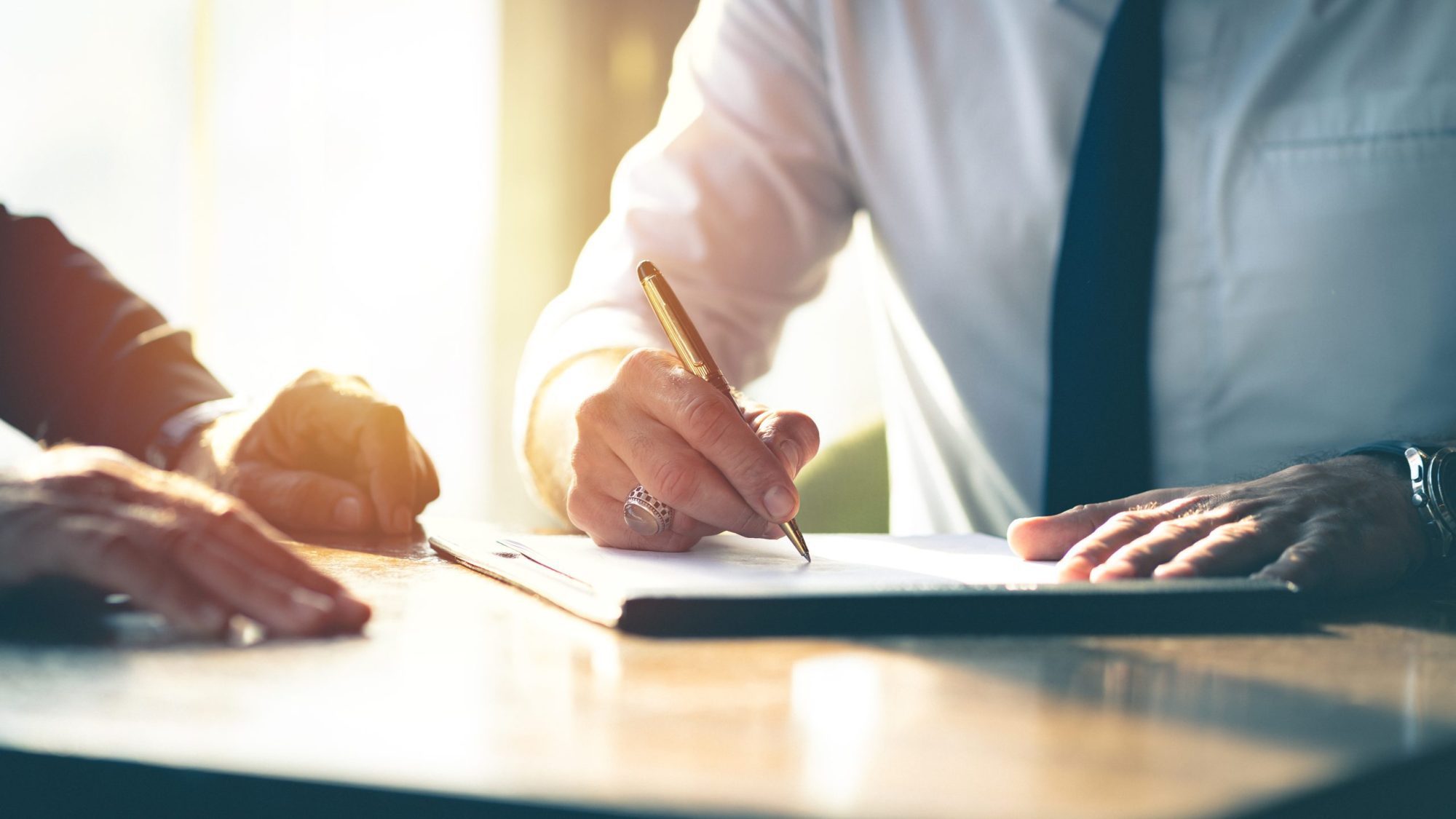Closeup Businessman signing a contract investment professional d Two people sit at a desk; one is signing a document with a pen while the other rests a hand on the table.