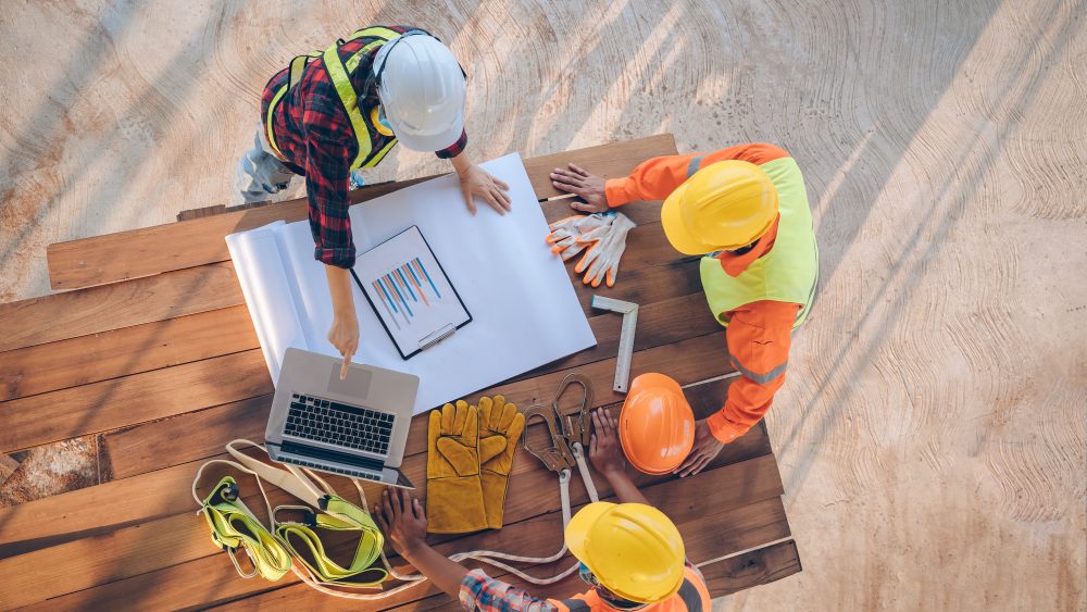 Three construction workers in safety gear review plans and a chart on a laptop at a wooden table with helmets and gloves on top.