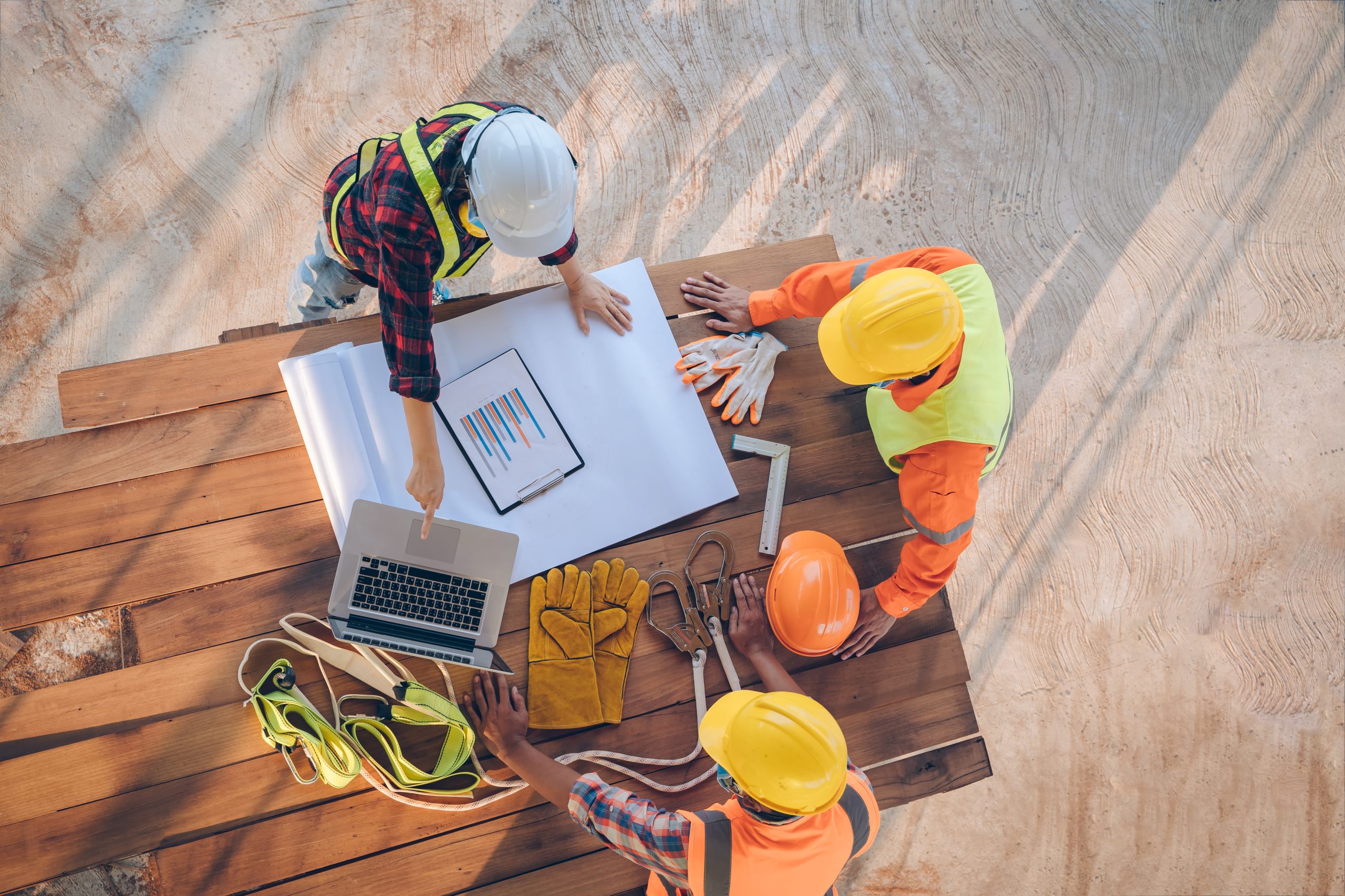 Team of engineer and architects working, meeting, discussing,des Three construction workers in safety gear review plans and a chart on a laptop at a wooden table with helmets and gloves on top.
